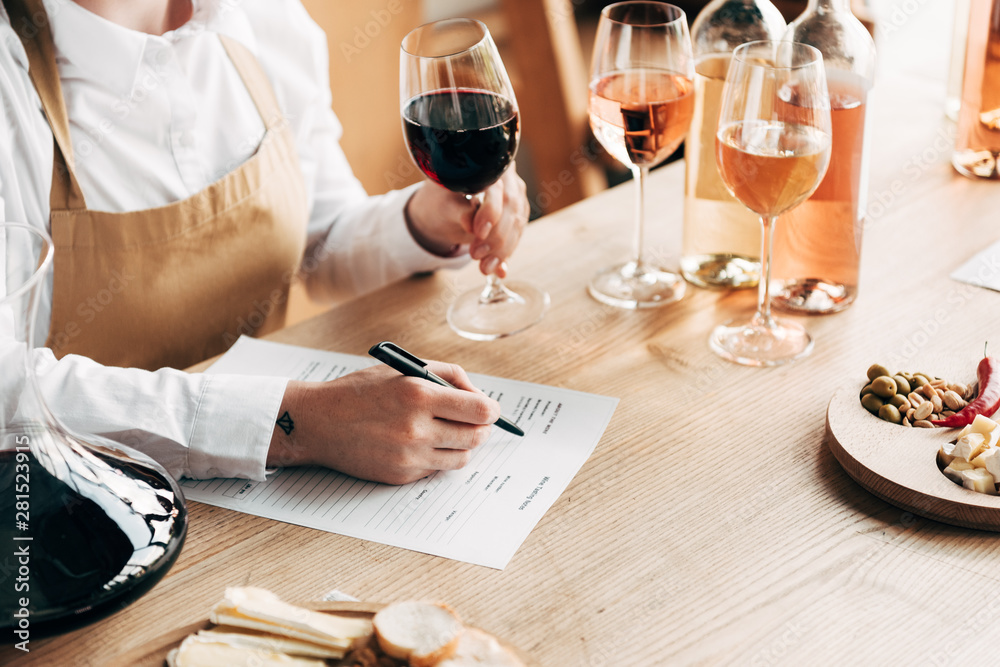 cropped view of sommelier in apron sitting at table, holding wine glass and writing in wine tasting document - obrazy, fototapety, plakaty 