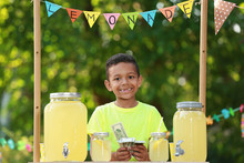 Cute Little African-American Boy With Money At Lemonade Stand In Park. Summer Refreshing Natural Drink
