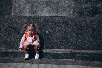 Wall Mural - little pretty baby who got lost in the city, a girl in a pink raincoat, with a backpack and a stylish hairstyle sitting next to a large administrative building one by one