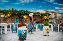 Young Romantic Woman Walks Near The Colorful Outdoor Cafe  In The Beautiful Sicilian Coastal Village Marzamemi In Sicily, South Italy
