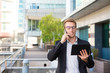 Cheerful manager talking on phone and reading on tablet screen outside. Young business man standing near office building and using digital gadgets. Communication concept