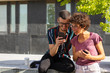 Happy nerdy couple watching funny content on phone together. Man and woman in glasses sitting on parapet outside, staring at phone screen and talking. Communication concept
