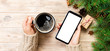 © sosiukin - Female hands holding modern smartphone with mosk up and mug of coffee on wooden table with christmas decoration. banner top view