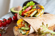 A healthy lunch or dinner of a vegan / vegetarian wrap made with  argula lettuce, sliced tomatoes, cucumbers, avocado, bell peppers and carrots. Selective focus on sandwich on top. 