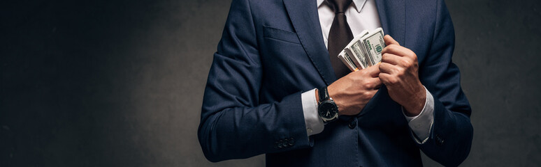 panoramic shot of businessman in suit putting cash in pocket on grey