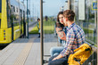© Iryna - Positive relax couple traveler drinking coffee outdoor at stop while waiting tram, sits on bench at sunny morning.