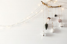Christmas Vintage Toys Hanging On Wooden Branch On White Wall With Festive Lights In Modern Room, Scandinavian Minimal Style. Stylish Glass Ornaments, Holiday Decorations. Space For Text