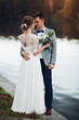 Back view of husband embracing his beautiful elegant wife in lace wedding dress with lovely bouquet against unfocused lake surface.