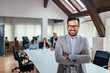 © bnenin - Smiling business leader looking at camera with arms crossed. Colleagues in the background.