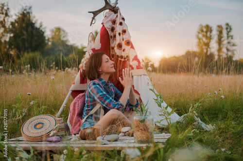 Plakat hipster woman in the form of a Shaman is looking for inspiration from mother Earth in a wigwam in nature.