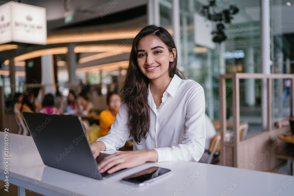 A young and attractive Indian Asian woman small business entrepreneur smiles as she works on her laptop in a cafe during the day. She is wearing a business casual outfit and is hopeful and confident. - obrazy, fototapety, plakaty 
