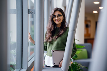 Portrait of a beautiful, young, elegant and attractive Indian Asian woman student in a preppy green sweater and spectacles. She is relaxing in a glass building during the day.