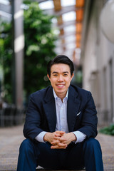 Wall Mural - Portrait of a young, confident and good-looking Chinese Asian business man in a well-fitted navy blue suit over a pale blue shirt. He is sitting on steps in the city and waiting for someone.