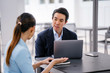 © Danon - A young, handsome and well-groomed Asian man in a dark suit is interviewing a professional woman candidate applying a job during the day. He is smiling as he talks to the woman.