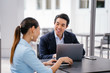 © Danon - A young, handsome and well-groomed Asian man in a dark suit is interviewing a professional woman candidate applying a job during the day. He is smiling as he talks to the woman.
