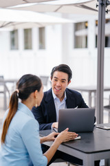 Wall Mural - A young, handsome and well-groomed Asian man in a dark suit is interviewing a professional woman candidate applying a job during the day. He is smiling as he talks to the woman.