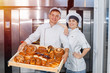 © salomonus_ - Bakers man and girl with a full box of hot pastries in their hands ha the background of an industrial oven in a bakery.