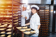 © salomonus_ - Baker girl with a baking sheet with raw dough in hands on the background of an industrial oven in a bakery.