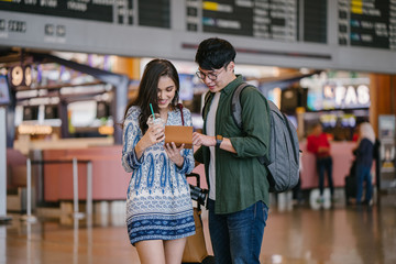 Wall Mural - Portrait of a young Asian interracial couple (Korean man and his Indian girlfriend) checking their passports and tickets in an airport. They are smiling excitedly for their holiday trip / vacation.