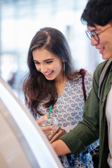 Wall Mural - A portrait of a pair of young and excited Asian travelers (Korean man and his Indian friend) checking in to board their plane for their holiday at an automated check-in booth in the airport.