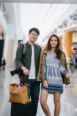 Wall Mural - Portrait of a young, attractive and diverse Asian couple (Korean man and his Indian girlfriend) are going for a holiday. They are casually dressed as stand in the middle of an airport during the day.