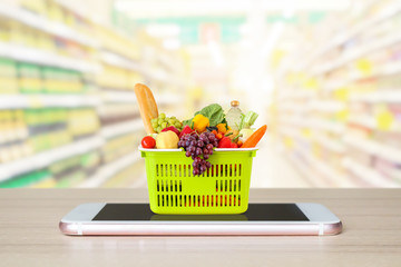 Wall Mural - Fresh food and vegetables in shopping basket on mobile smartphone on wood table with supermarket aisle blurred background grocery online concept