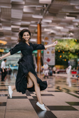 Wall Mural - Portrait of an attractive, young and athletic north Indian Asian woman smiling and laughing as she leaps and jumps indoors in a mall or airport. She is wearing a casual black flowing dress.