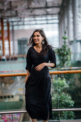 Wall Mural - Portrait of a gorgeous, beautiful, tall and elegant Indian Asian girl in a black casual dress smiling in a modern mall or airport during the day.