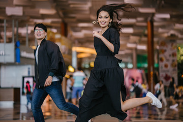 A diverse Asian pair (Indian woman and a Korean man) smile and laugh as they leap and jump together in sync in a modern airport during the day. They are casually dressed and are having great fun.