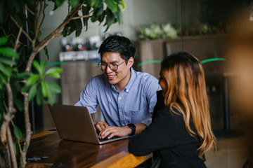 A pair of diverse team mates have a business meeting and discussion together to collaborate. A Korean man is sitting at a table in a trendy coworking space office and talking to his female companion.