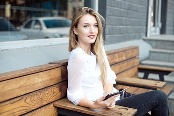 Attractive young woman in vintage jeans sitting on a chair in an outdoor restaurant. Attractive blonde in a white shirt is waiting for an order in her favorite cafe and enjoys nature