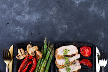 Wall Mural - Grilled chicken breast on a cast iron skillet with grill vegetables on a stone background, flat lay