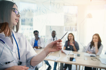 African professor at medical conference in modern clinic teaches his students. Doctor writing on board some formulas for interns in conference room at hospital at sunrise.