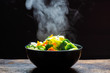 canvas print picture - The steam from the vegetables carrot broccoli Cauliflower in a black bowl, a steaming. Boiled hot Healthy food on table on black background,hot food and healthy meal concept