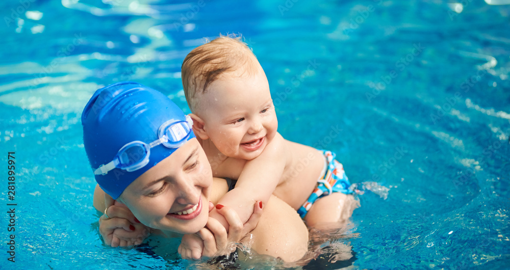 Child having fun in water with mom. Little wet boy smiling holding on his mother's back and happy woman with swimming cap and goggles. Blue turquoise water on background. Horizontal portrait view - obrazy, fototapety, plakaty 