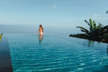 Slim young woman in swimsuit relaxing on edge tropical infinity pool in mountains. Palms around and crystal clean water. Luxury resort on Bali island