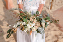 Bride With Wedding Bouquet Is On Beach Near Blue Sea After Ceremony. Bridal Dress Is White, Lace, Boho Style