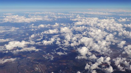 Wall Mural - Flying over beautiful heaps of white clouds moving softly from right to left on the bright blue sky, and countryside landscapes far beneath. Picturesque view from the airplane window.