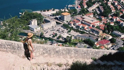 Wall Mural - Rear view of young woman in dress and hat, who looking from above to the Kotor city and Boka bay, Montenegro