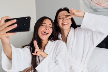 Wall Mural - Young pretty smiling women in white bathrobes happily showing two fingers gesture taking cool photos together on cellphone at home