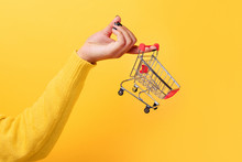 Buying Things At Market Shops Concept. Woman Hand Holding Small Tiny Shopping Cart Trolley Over Trend Yellow Background