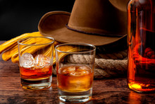 The Wild West Concept Theme With Cowboy Hat, Rope Lasso, Leather Gloves, Two Glasses Of Whiskey On The Rocks And Bottle Of Bourbon On Wooden Table In A Vintage Saloon Against Black Background