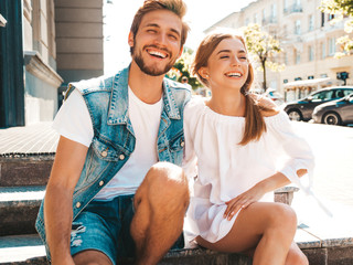 Smiling beautiful girl and her handsome boyfriend. Woman in casual summer dress and man in jeans clothes. Happy cheerful family. Sitting on stairs on the street background.Hugging couple