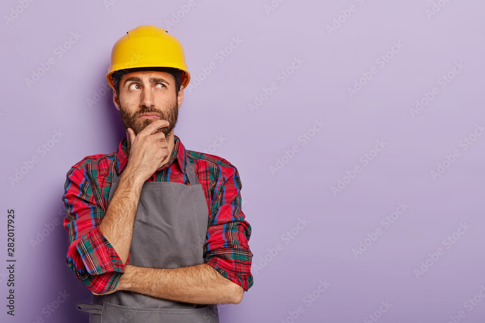 Thoughtful man builder construction touches chin, thinks over new idea for building, works as repairman, wears yellow protective helmet, checkered shirt and apron. Industry and repairing concept - obrazy, fototapety, plakaty 