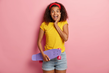 Happy teenage girl with Afro hair holds skateboard, has fun with friend while skating, enjoys active rest, wears yellow t shirt and denim shorts, poses over pink background. People, hobby, leisure