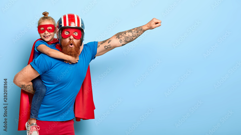 Horizontal shot of powerful man makes fly gesture, gives piggyback to small daughter dressed in superhero suit, have supernatural abilities, enjoy free time together. Real heroes defend you. - obrazy, fototapety, plakaty 