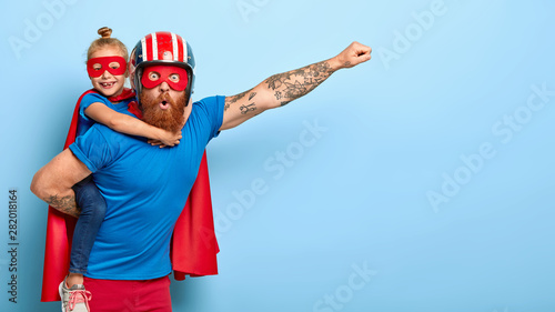 Fotomural Horizontal shot of powerful man makes fly gesture, gives piggyback to small daughter dressed in superhero suit, have supernatural abilities, enjoy free time together