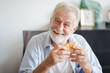 Seniors European man is sitting to eat a burger at home which looking at camera. Retired man is smiling with felling happy and take a burger on hand.