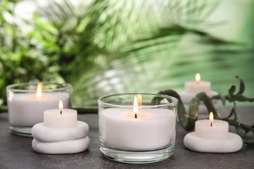 Burning candles and spa stones on grey table against blurred green background, space for text