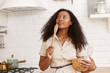 © Anatoliy Karlyuk - Indoor shot of attractive young dark-skinned woman in beige dress posing in kitchen interior holding wooden spoon, bowl and whisk going to make dough for pancakes, looking up with inspired smile